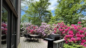 An outdoor deck with a wooden table and chairs, surrounded by blooming pink and white flowers. - Family Home with Fire Pit, Games & Pool Access (Somerset)