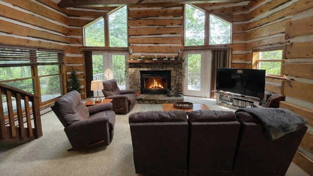 A log cabin interior with a fireplace, leather sofas, a TV, and large windows. - Family Home with Fire Pit, Games & Pool Access (Somerset)