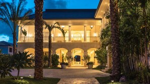 Un edificio de dos pisos con una entrada central, rodeado de palmeras y un camino pavimentado que conduce a la entrada.