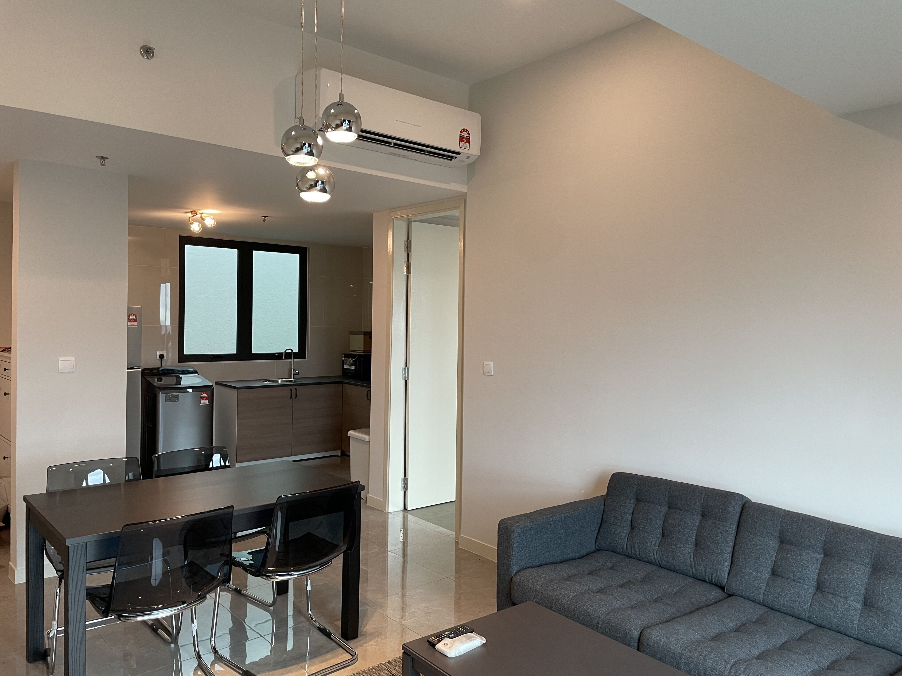 Una sala de estar moderna con un sofá gris, una mesa de comedor de vidrio con sillas y una cocina con ventana y electrodomésticos integrados.
