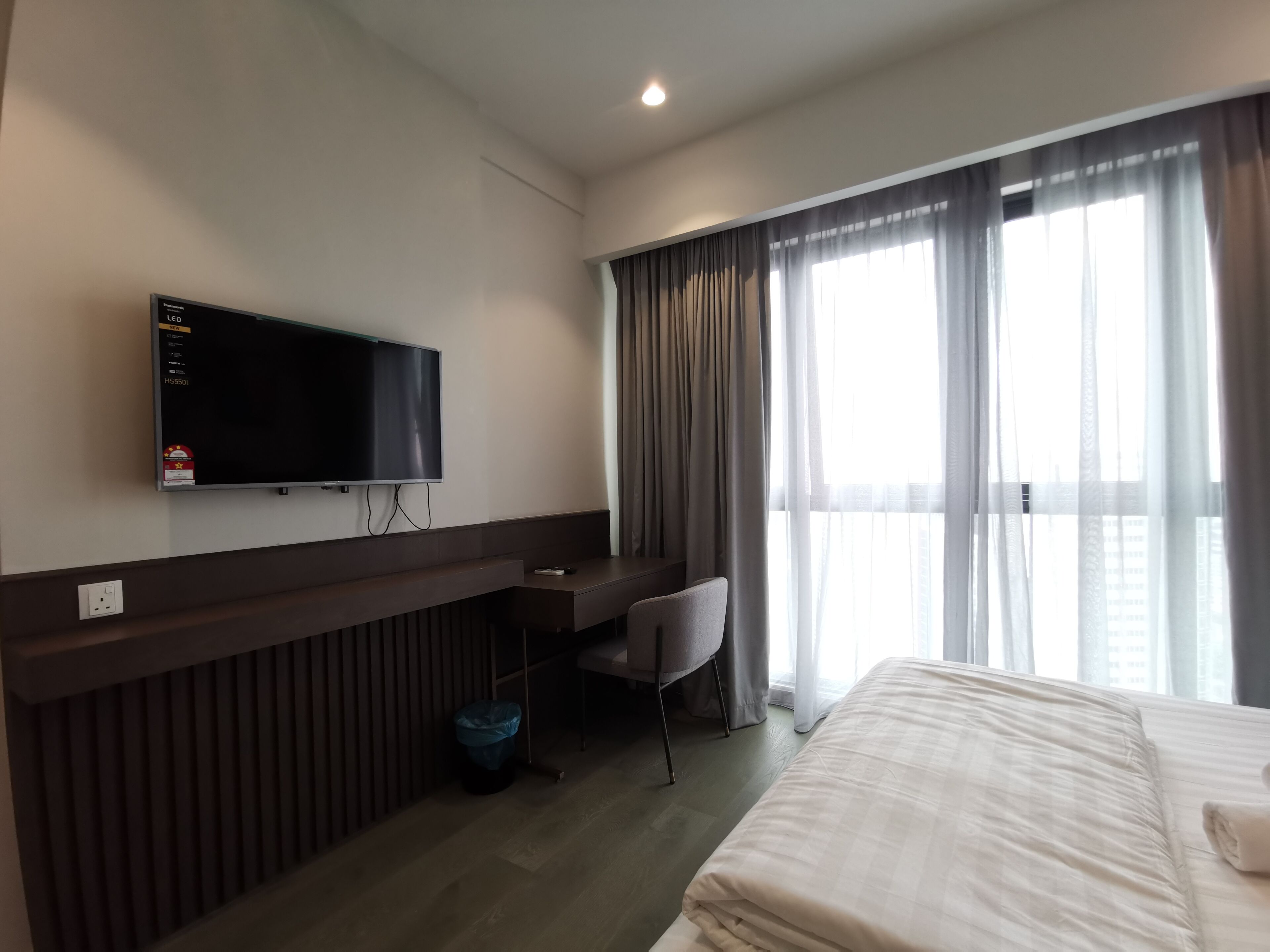 Une chambre d’hôtel comprenant un lit, un bureau avec une chaise, un téléviseur fixé au mur et une fenêtre avec des rideaux.