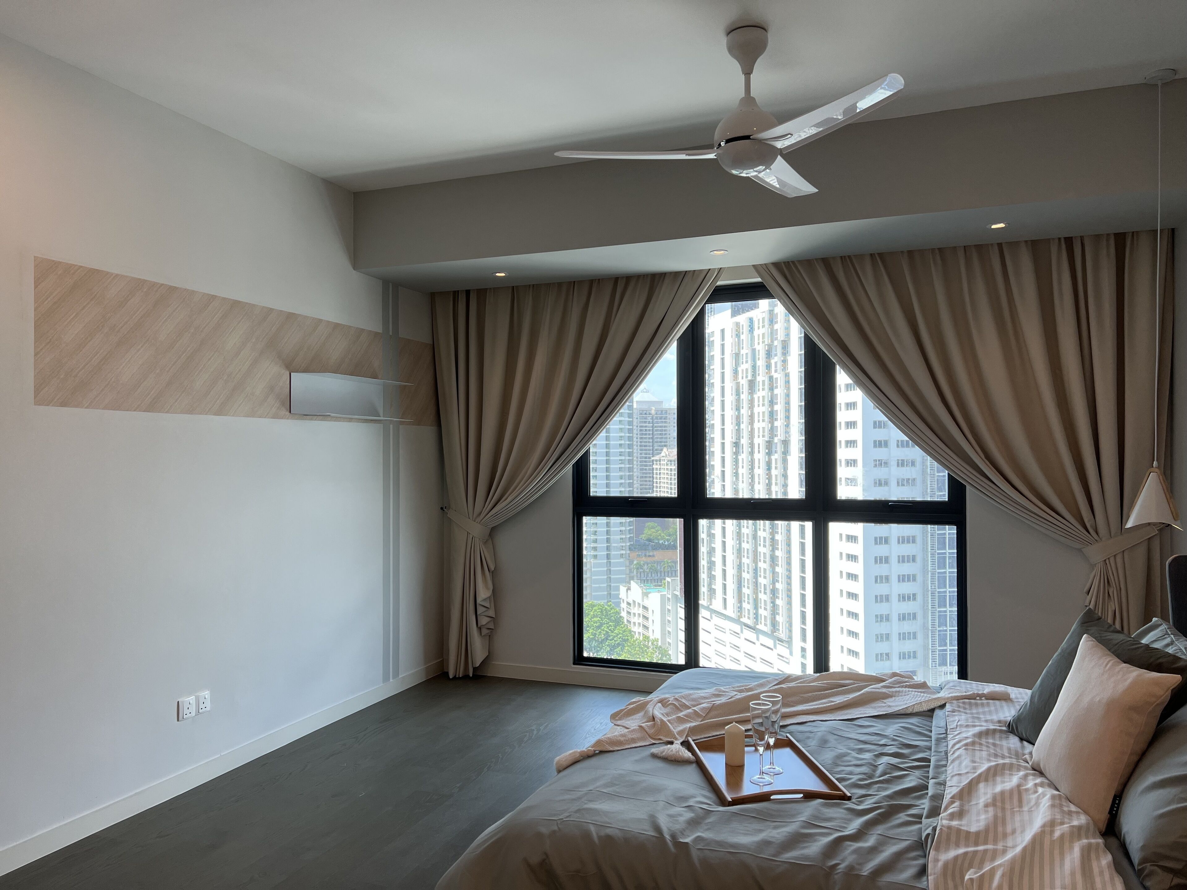 Une chambre d’hôtel moderne avec un grand lit, un ventilateur de plafond et une vue sur les grands immeubles à travers de larges fenêtres.
