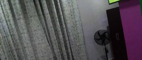 A hotel room with a bed, curtains, a TV on the wall, and a fan.