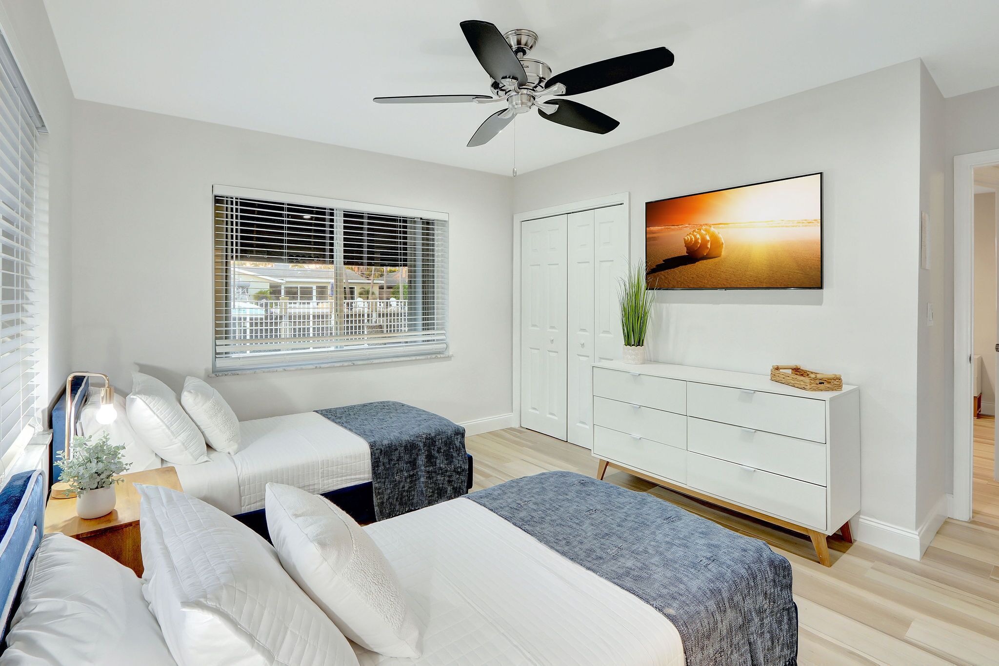 A modern bedroom with two beds, a ceiling fan, a large TV, and a window with blinds.