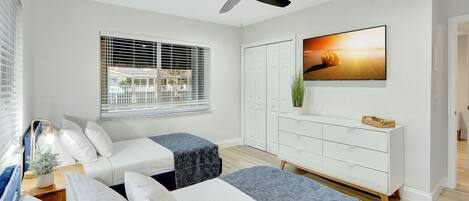 A modern bedroom with two beds, a ceiling fan, a large TV, and a window with blinds.