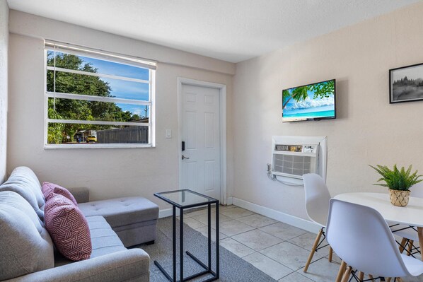 A modern living room with a sofa, a glass coffee table, a TV mounted on the wall, and a dining area with a chair and table.