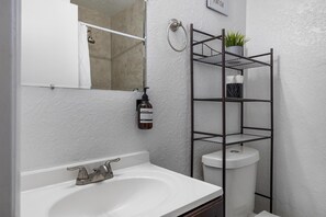 A bathroom with a white sink, a toilet, a shower, and a metal shelf unit with a plant.