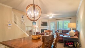 Un comedor con mesa y sillas de madera, una araña, una escalera, una sala de estar con sofá y televisor.