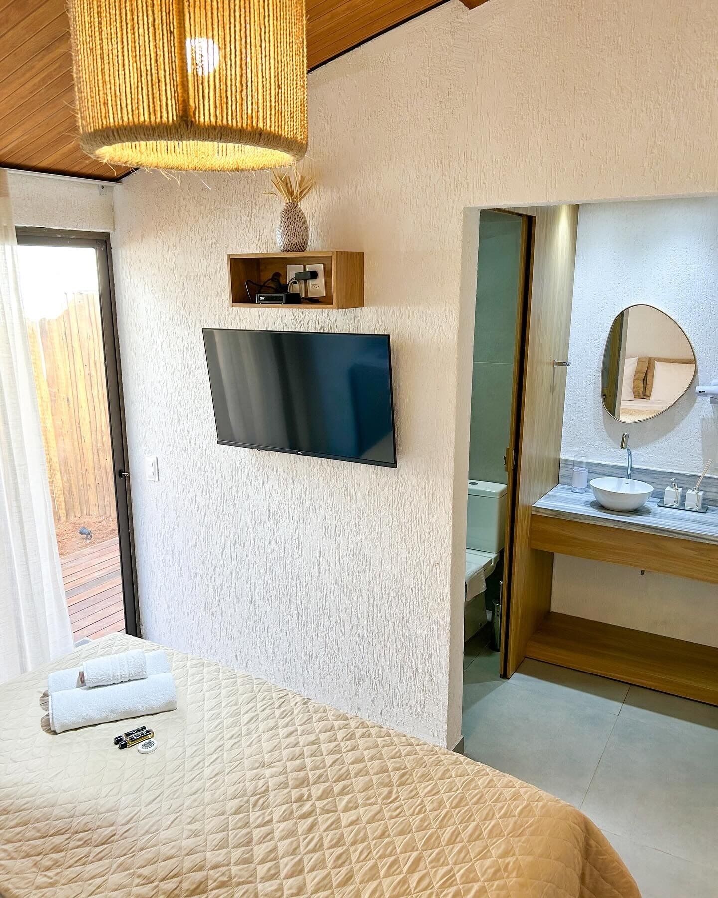A bedroom with a bed, a television mounted on the wall, a wooden shelf with a vase, and a bathroom with a sink and mirror.