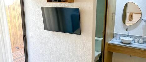 A bedroom with a bed, a television mounted on the wall, a wooden shelf with a vase, and a bathroom with a sink and mirror.
