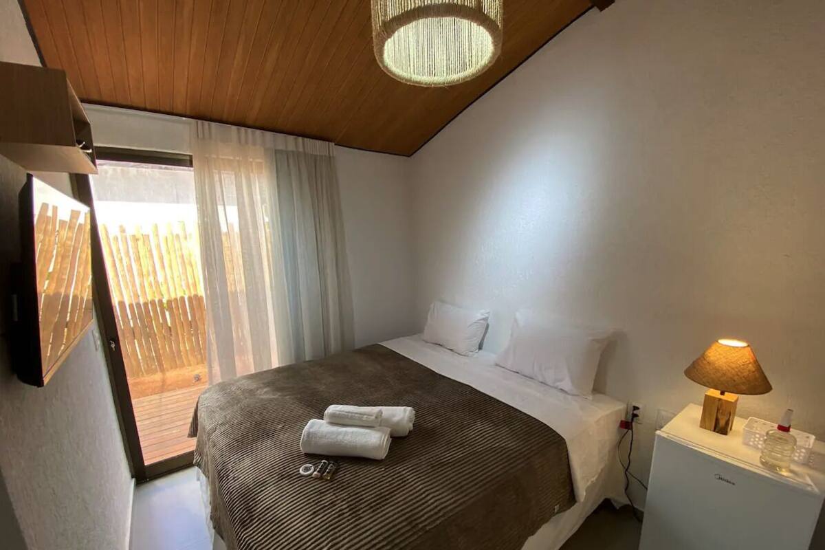 A bedroom with a bed, a nightstand, a lamp, and a view of a wooden deck through a window.
