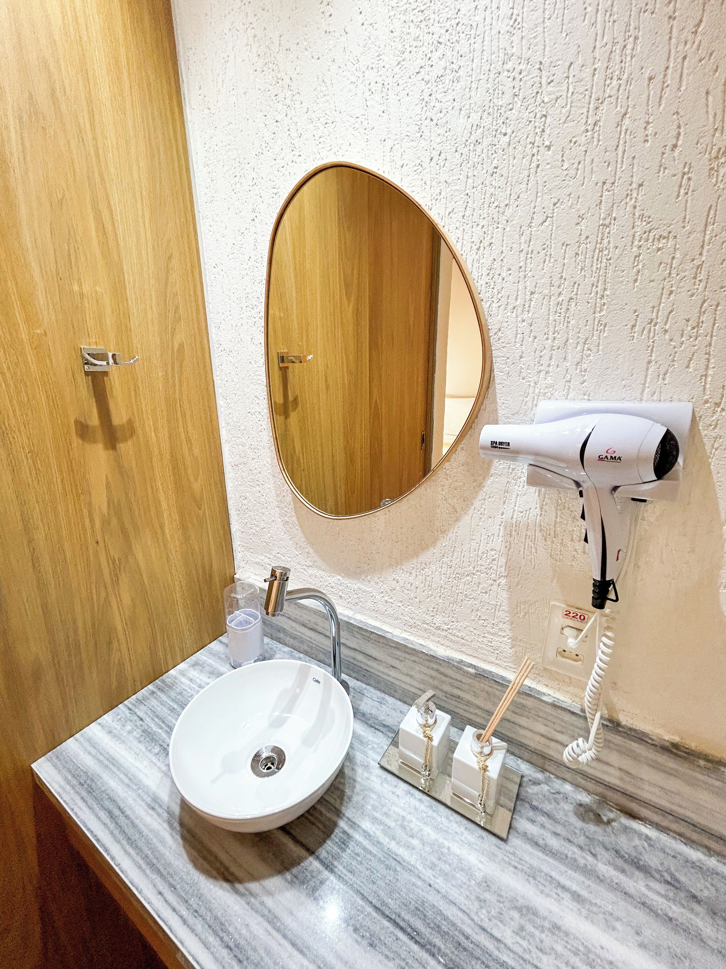 A bathroom with a wooden door, a round mirror, a sink with a marble countertop, a hairdryer, and a soap dispenser.