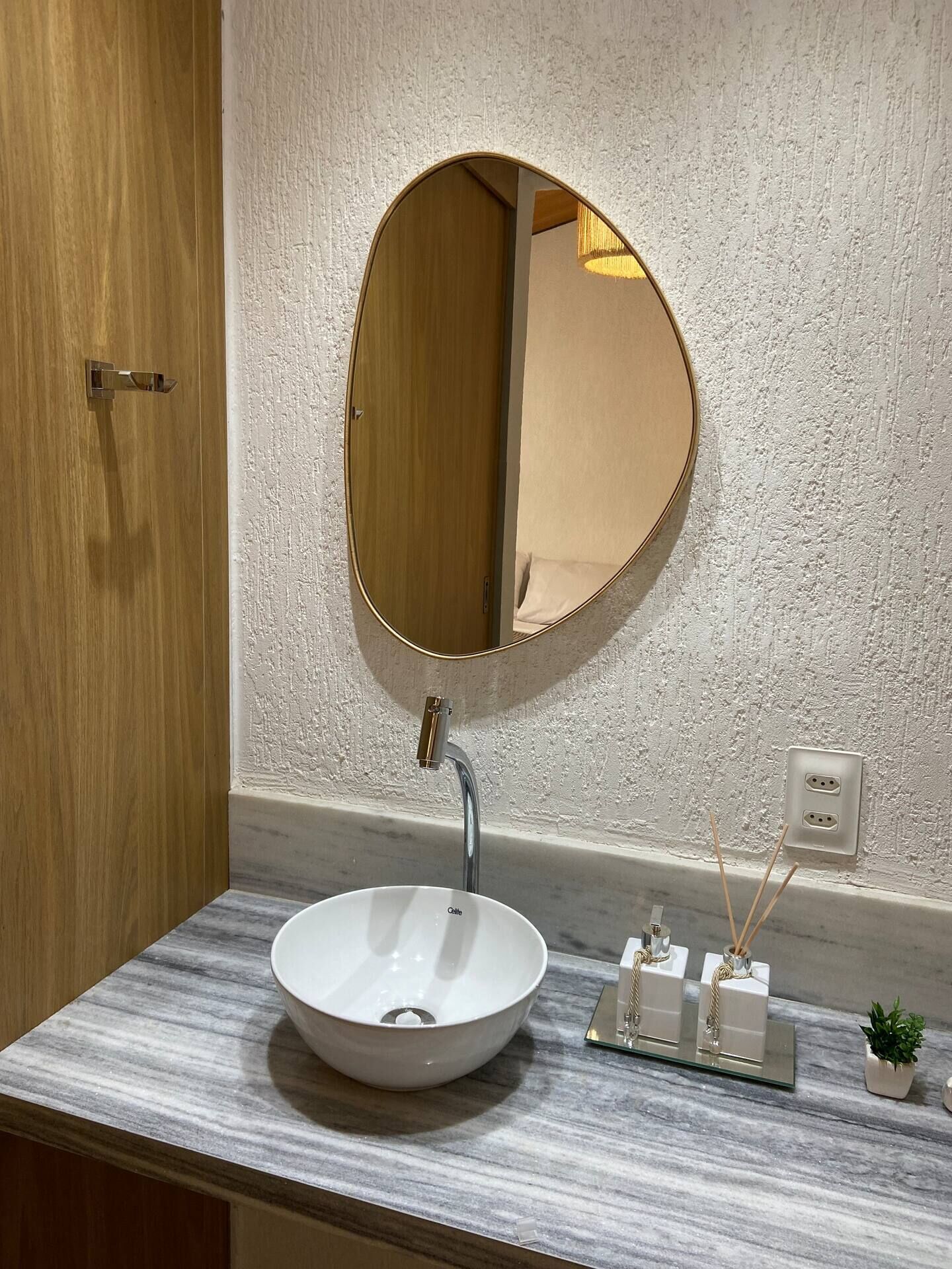 Un baño con un espejo redondo, un lavabo blanco, encimera de mármol, una pequeña planta en maceta y un enchufe en la pared.