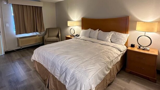 Habitación de hotel con una cama grande, dos mesitas de noche con lámparas, una silla, una ventana con cortinas y un aire acondicionado de pared.