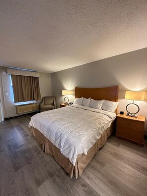 A hotel room with a large bed, two bedside tables with lamps, a chair, a window with curtains, and a wall-mounted air conditioner.