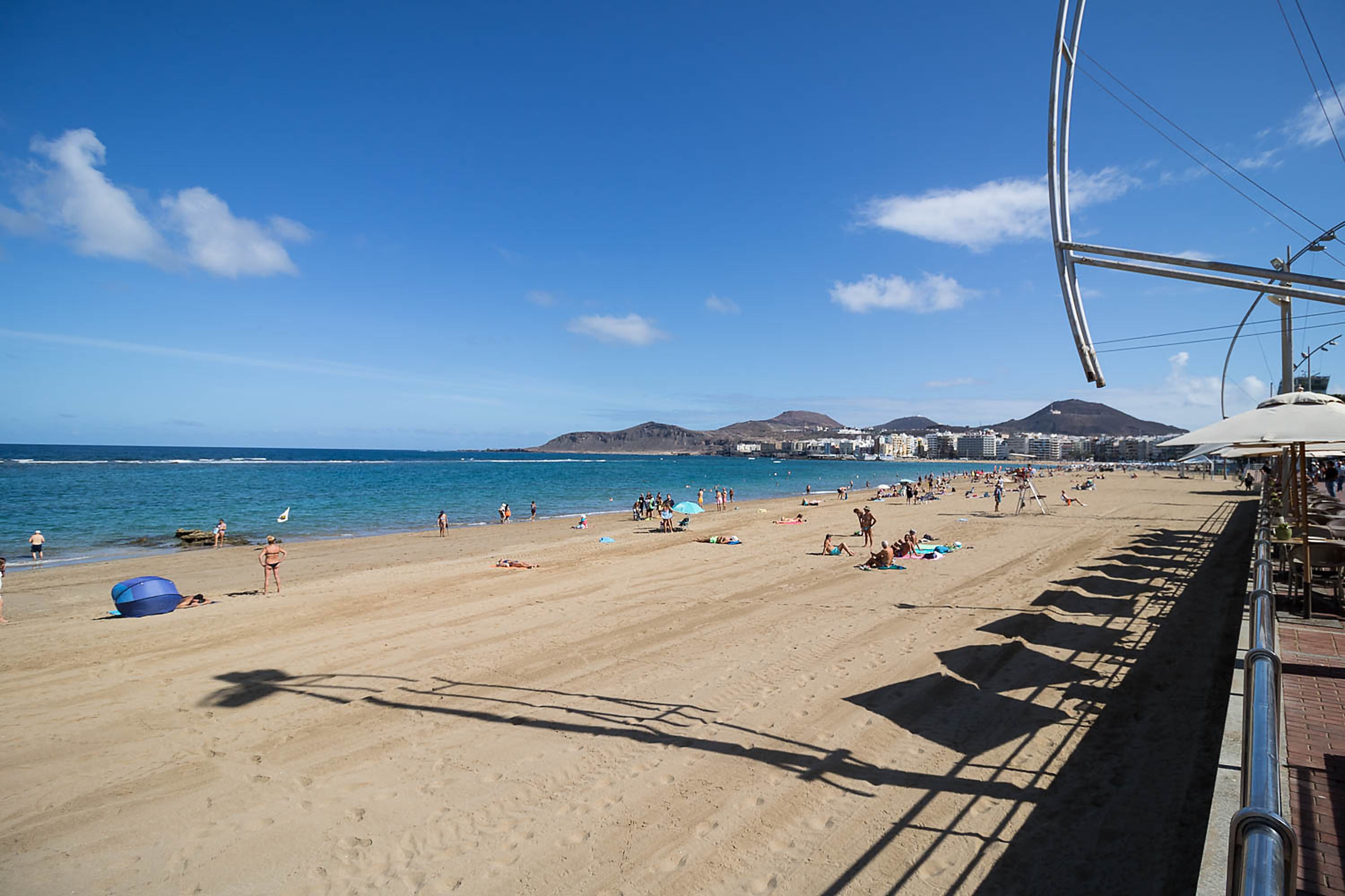Una playa con cielo despejado, orilla arenosa y un paisaje urbano al fondo.