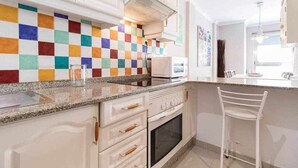 A kitchen with white cabinets, a colorful checkered backsplash, a granite countertop, and a white chair. - Oli Coral Canteras (Palmas De Gran Canaria, Las)