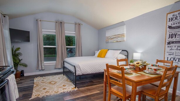 A bedroom with a bed, a dining table with chairs, a TV mounted on the wall, and a window with curtains. - Downtown Accessible New 1BR 1BA Cozy Studio (San Antonio)