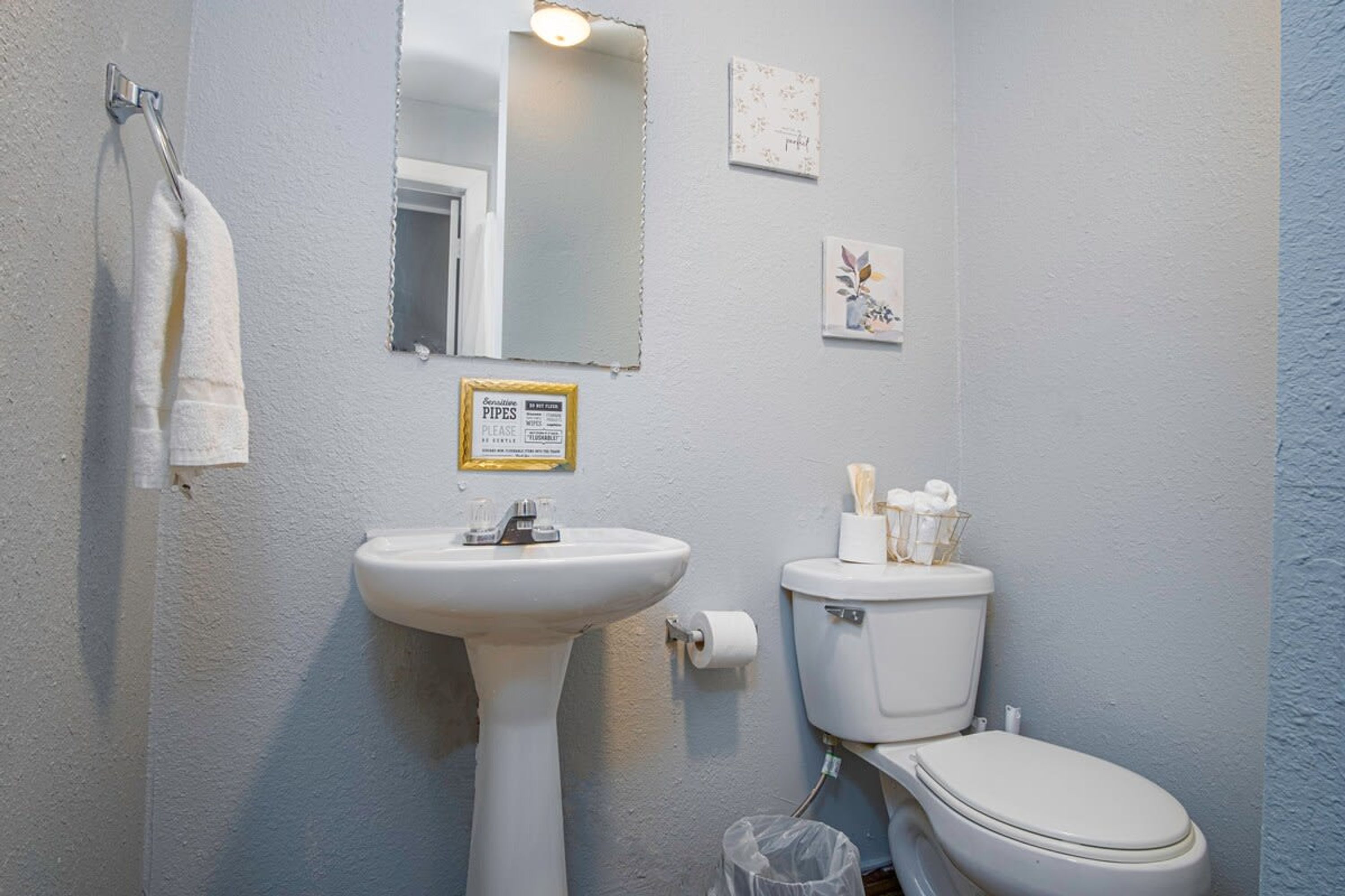 A bathroom with a white sink, toilet, and a towel hanging on a rack. There is a framed sign on the wall and a small picture.