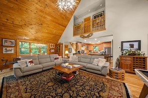 A spacious living area with a high ceiling, wooden beams, a large sofa set, a coffee table, and a view of the kitchen.