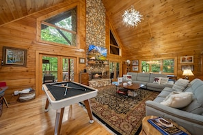 A wooden cabin interior with a pool table, a stone fireplace, a sofa, and a large window.