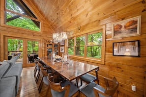 A wooden cabin interior with a large dining table, chairs, a sofa, and large windows offering a view of the forest.