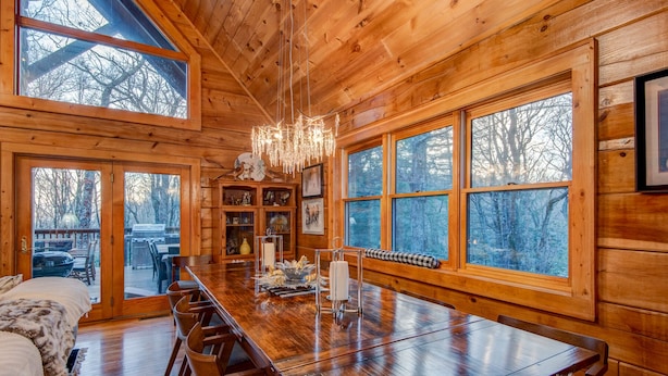 Interior de una cabaña de madera con una amplia mesa de comedor, varias sillas, un candelabro y grandes ventanales que ofrecen vistas del bosque circundante.