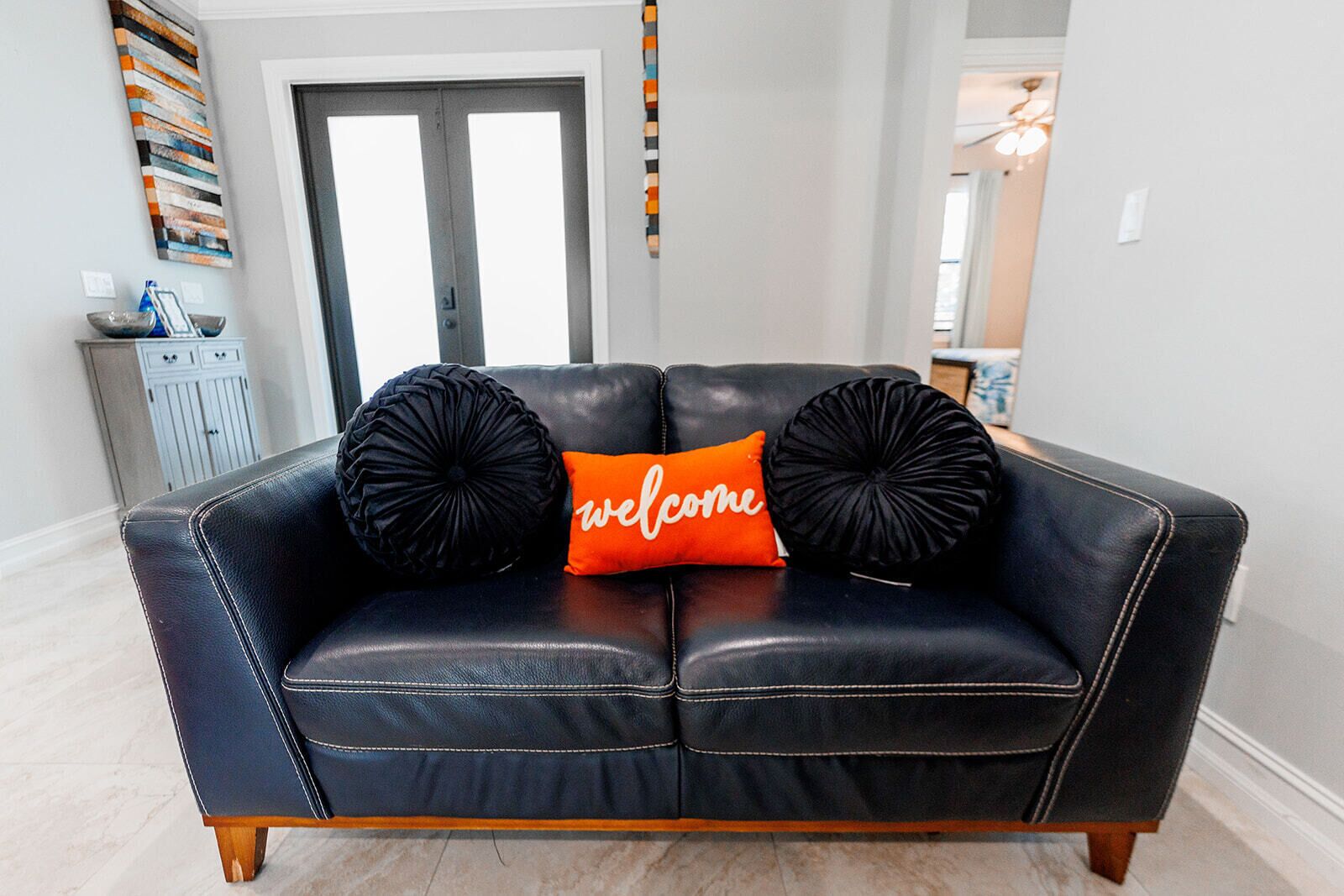 Un canapé en cuir noir, agrémenté de deux coussins décoratifs noirs et d’un seul coussin orange sur lequel est inscrit le mot « bienvenue », est placé dans une pièce dont le sol est de couleur claire et où l’on aperçoit une porte au fond.