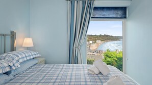 Et soveværelse med seng, natbord, lampe og udsigt til strand og hav.