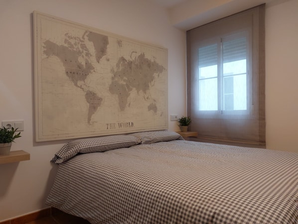 A bedroom with a large world map on the wall, a checkered bedspread, a window with blinds, and a small plant on a nightstand.