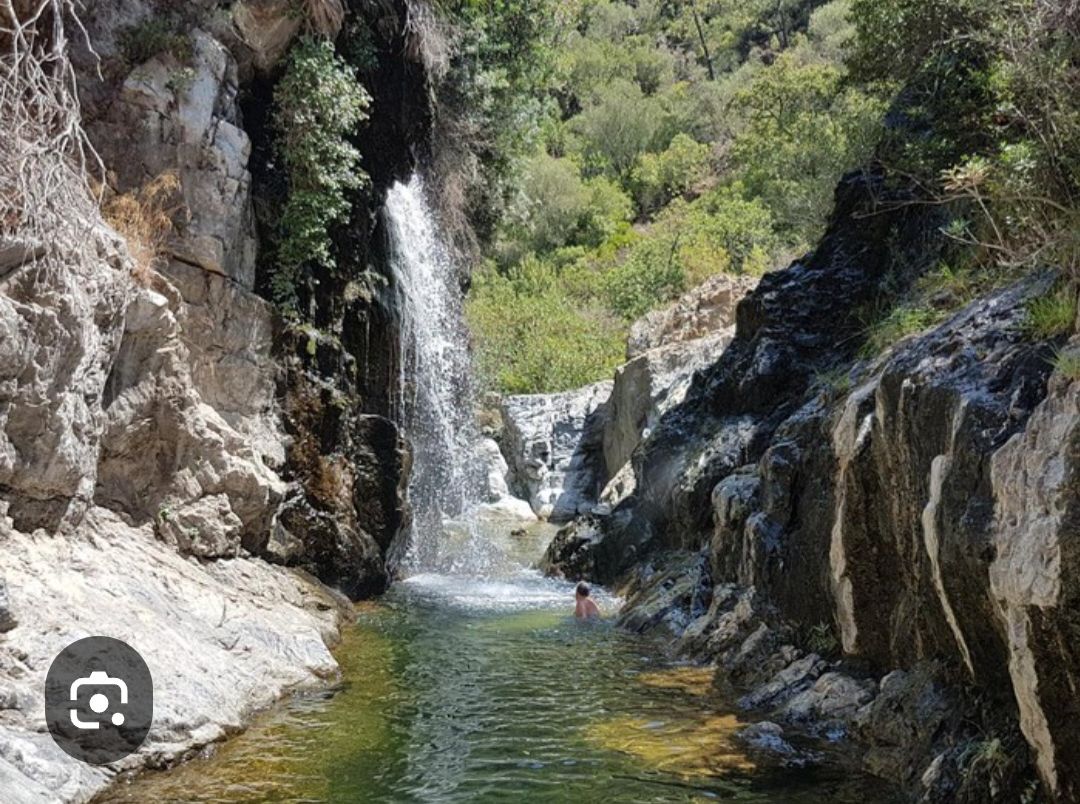 A natural hot spring with a waterfall, surrounded by rocky terrain and greenery.