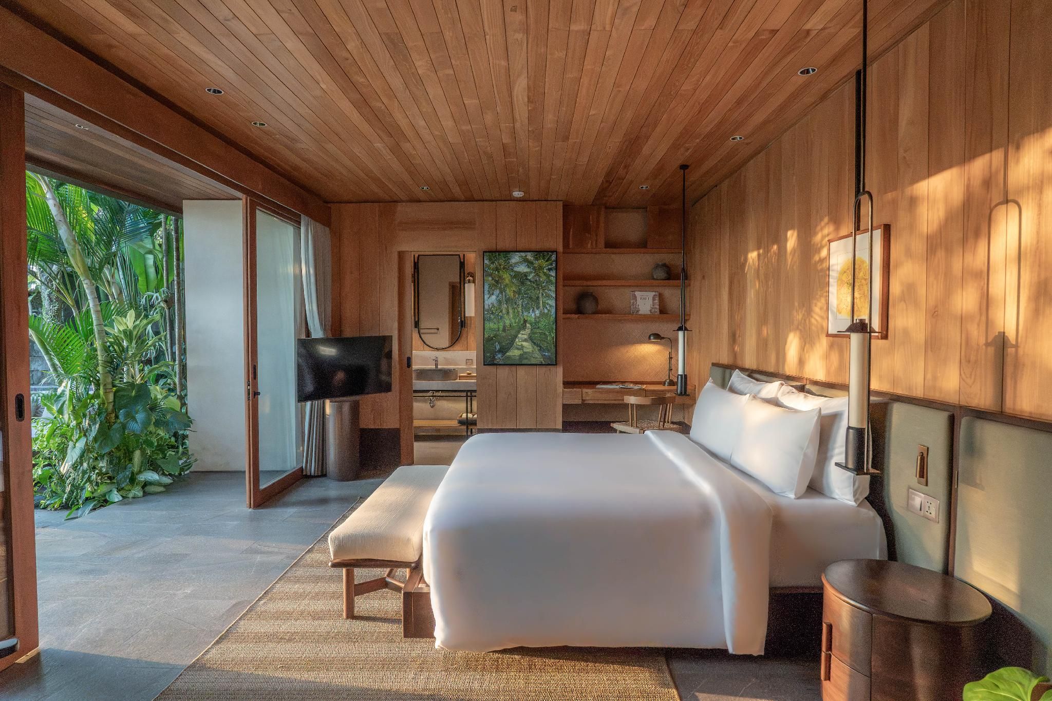 A modern bedroom with a large bed, a television, and a view of lush greenery outside.