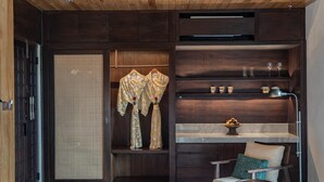 A modern interior with a wooden ceiling, a wardrobe with hanging clothes, a seating area with a chair and a table, and a shelf with decorative items. - The Ridge Bali (Ubud)