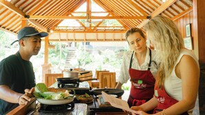 A group of people in a kitchen area with wooden beams, a ceiling fan, and a view of greenery outside. - The Ridge Bali (Ubud)