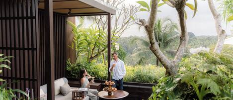 A wooden deck with a sofa and a table, surrounded by lush greenery and a view of the landscape.