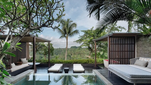 A modern outdoor pool area with lounge chairs and a sofa, surrounded by lush greenery and a view of the forest. - The Ridge Bali (Ubud)