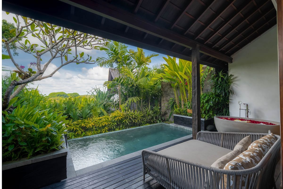 A private outdoor pool area with a wooden deck, wicker furniture, and lush tropical plants.