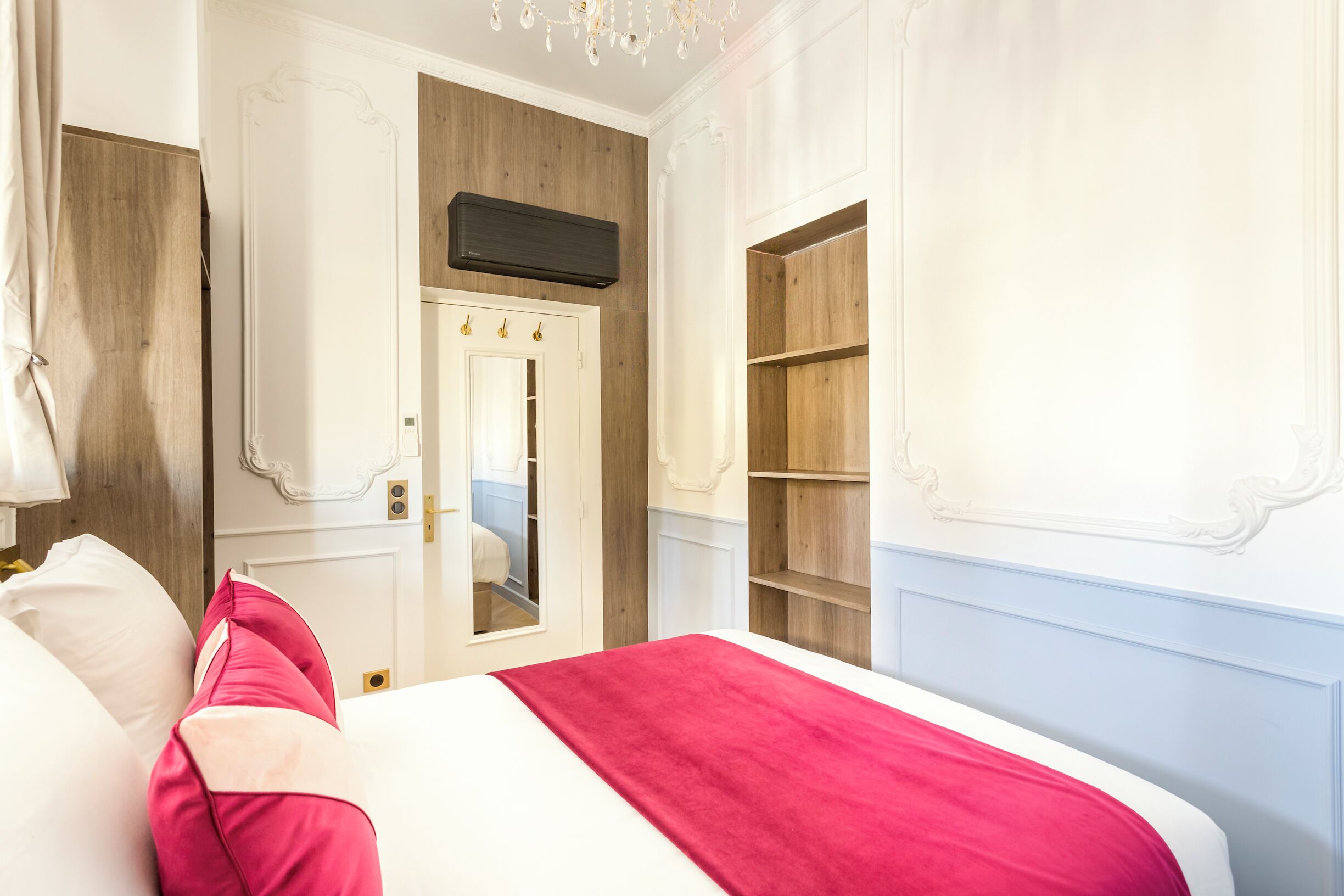 Quarto com uma cama, um cobertor vermelho, uma porta branca e prateleiras de madeira.