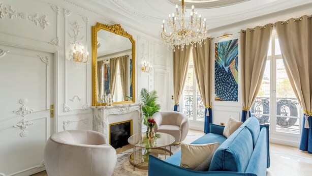 Una sala de estar lujosa con paredes blancas decoradas, una gran araña y un sofá azul, además de un espejo grande.