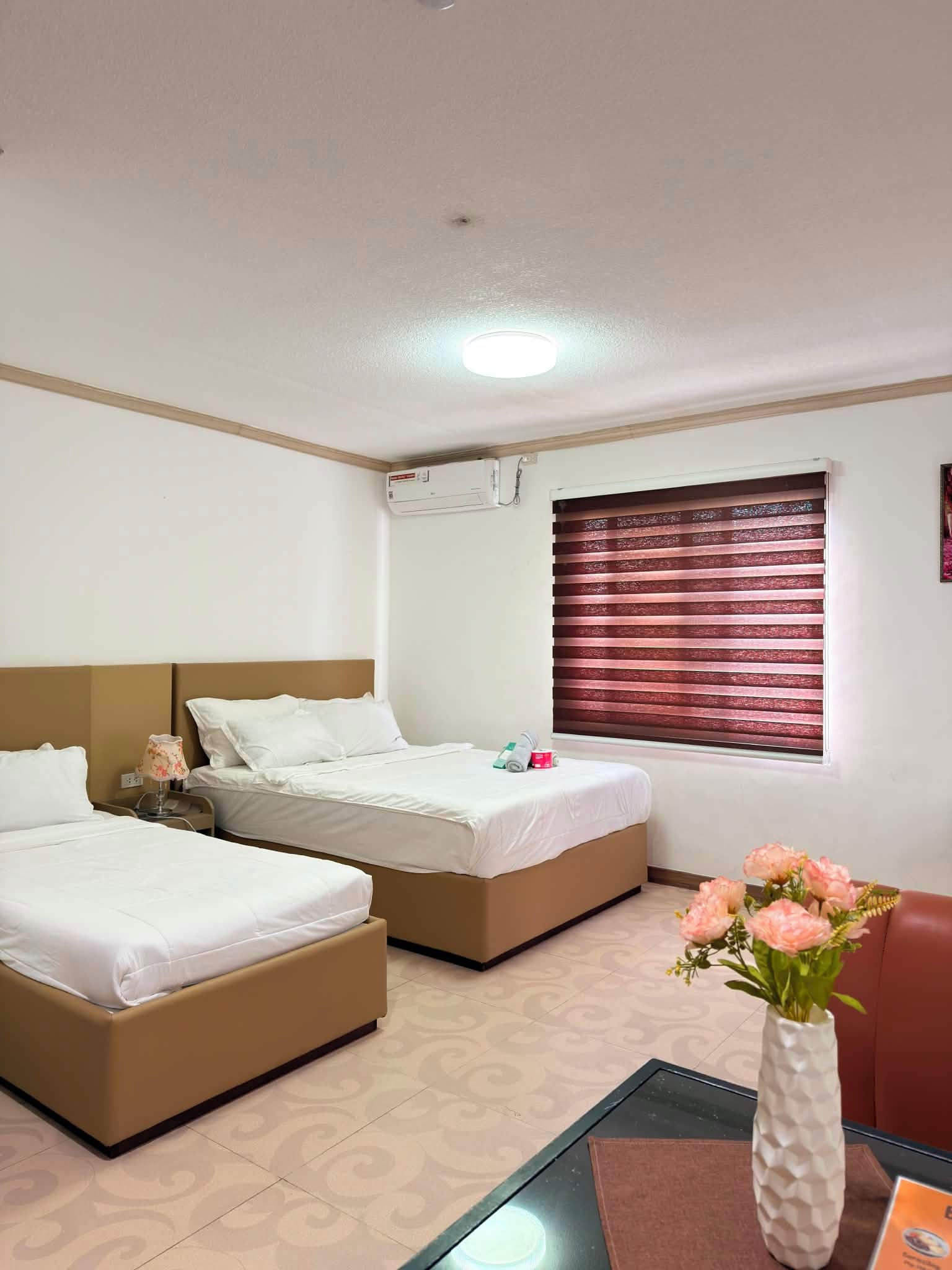 A hotel room with two beds, a window with blinds, a vase with flowers, and an air conditioning unit.