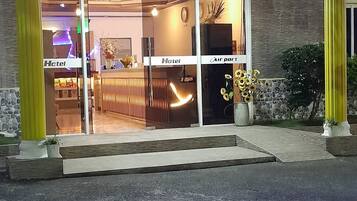 Entrada de hotel con un cartel luminoso, mostrador de recepción y señalización visible del aeropuerto.