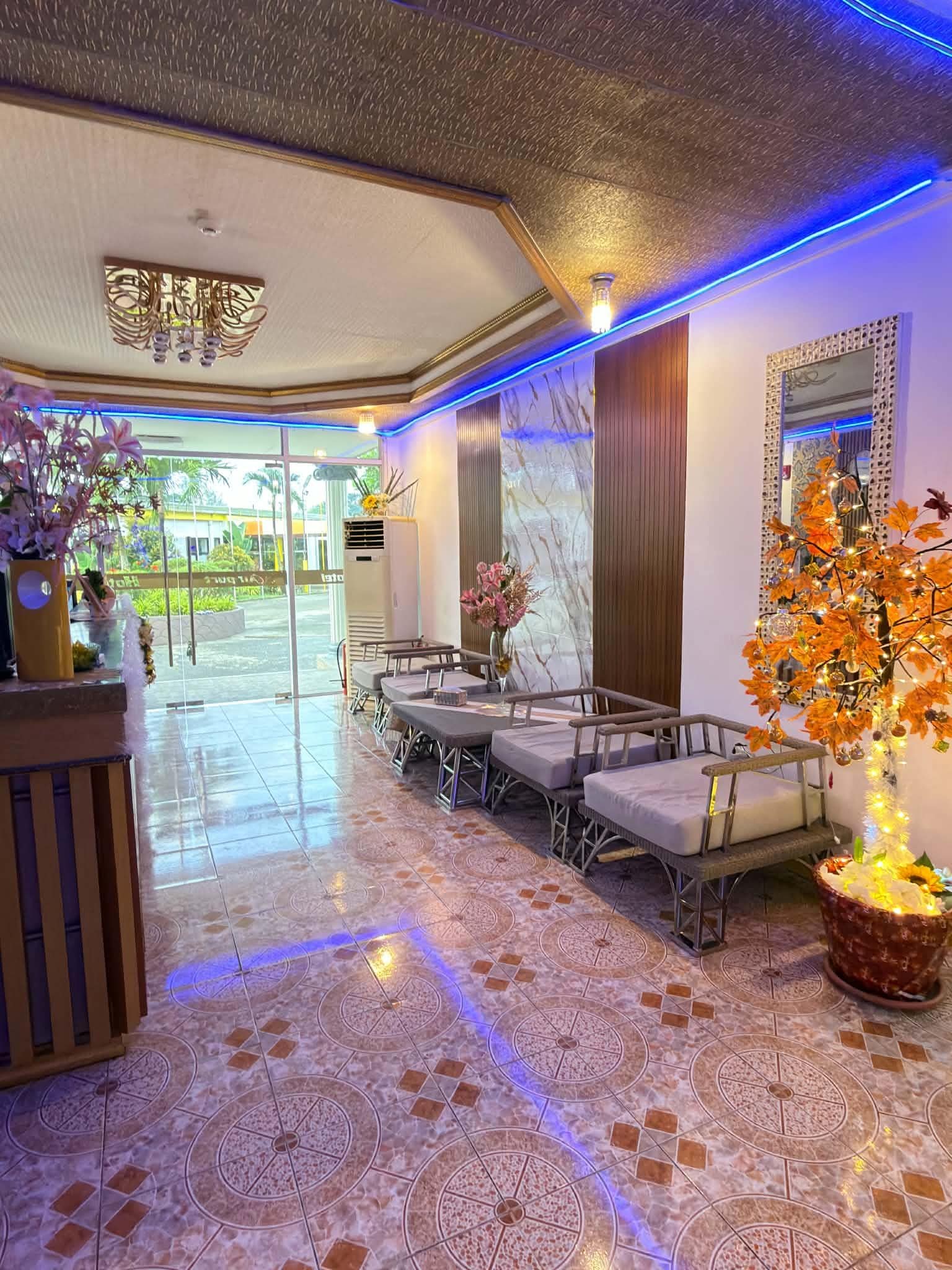 A hotel lobby with a reception desk, seating area, decorative plants, and a large mirror on the wall.