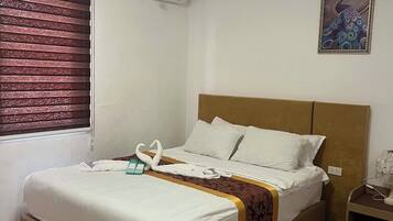 Ein Hotelzimmer mit Bett, Fenster mit Jalousien, Klimaanlage, Nachttisch und einem Wandbild.