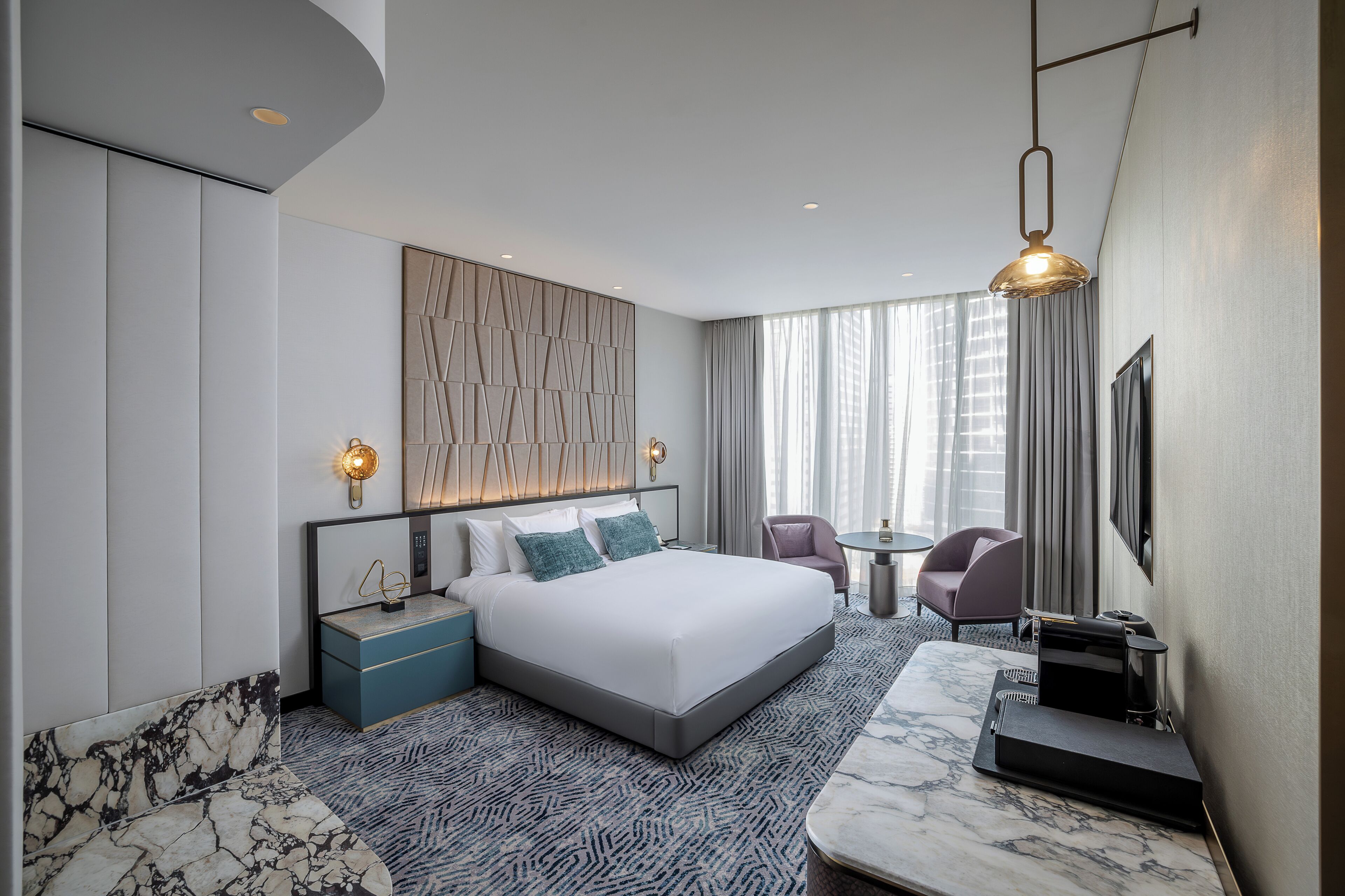 Quarto de hotel moderno com uma cama grande, área de estar com um banco de mármore, uma pequena mesa e uma televisão.