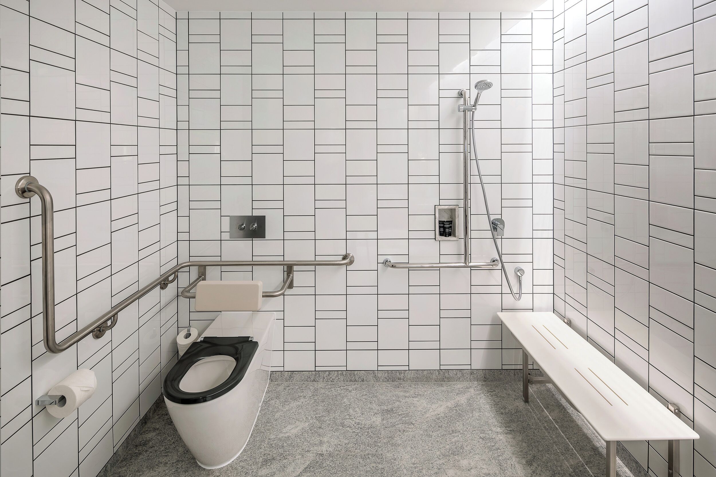 A small, modern bathroom with white tiled walls, a toilet, a bench, and a shower area.