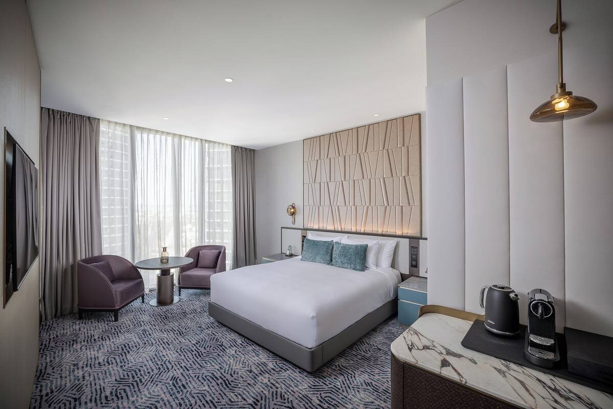 Ein modernes Hotelzimmer mit einem großen Bett, zwei Sesseln, einem kleinen runden Tisch, einer Kaffeemaschine und einem Nachttisch mit Marmorplatte.