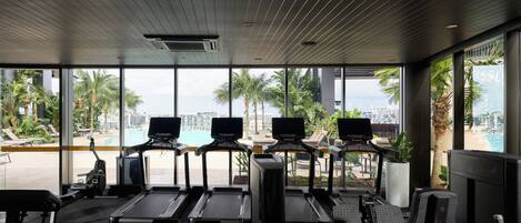 A gym with treadmills, exercise bikes, and a rowing machine, overlooking a pool and palm trees.