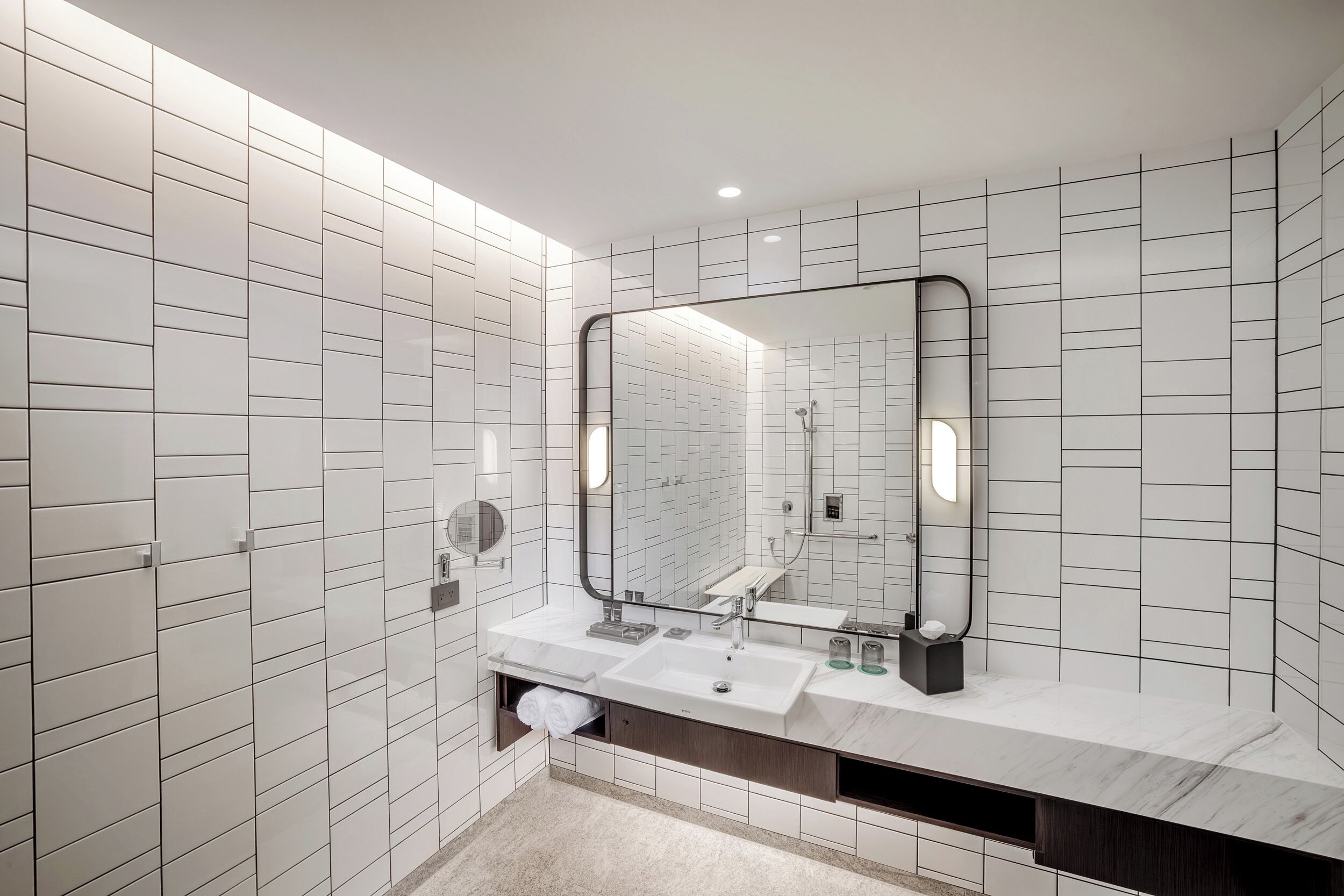 A modern bathroom with white tiled walls, a large mirror, a marble countertop, and a built-in sink.