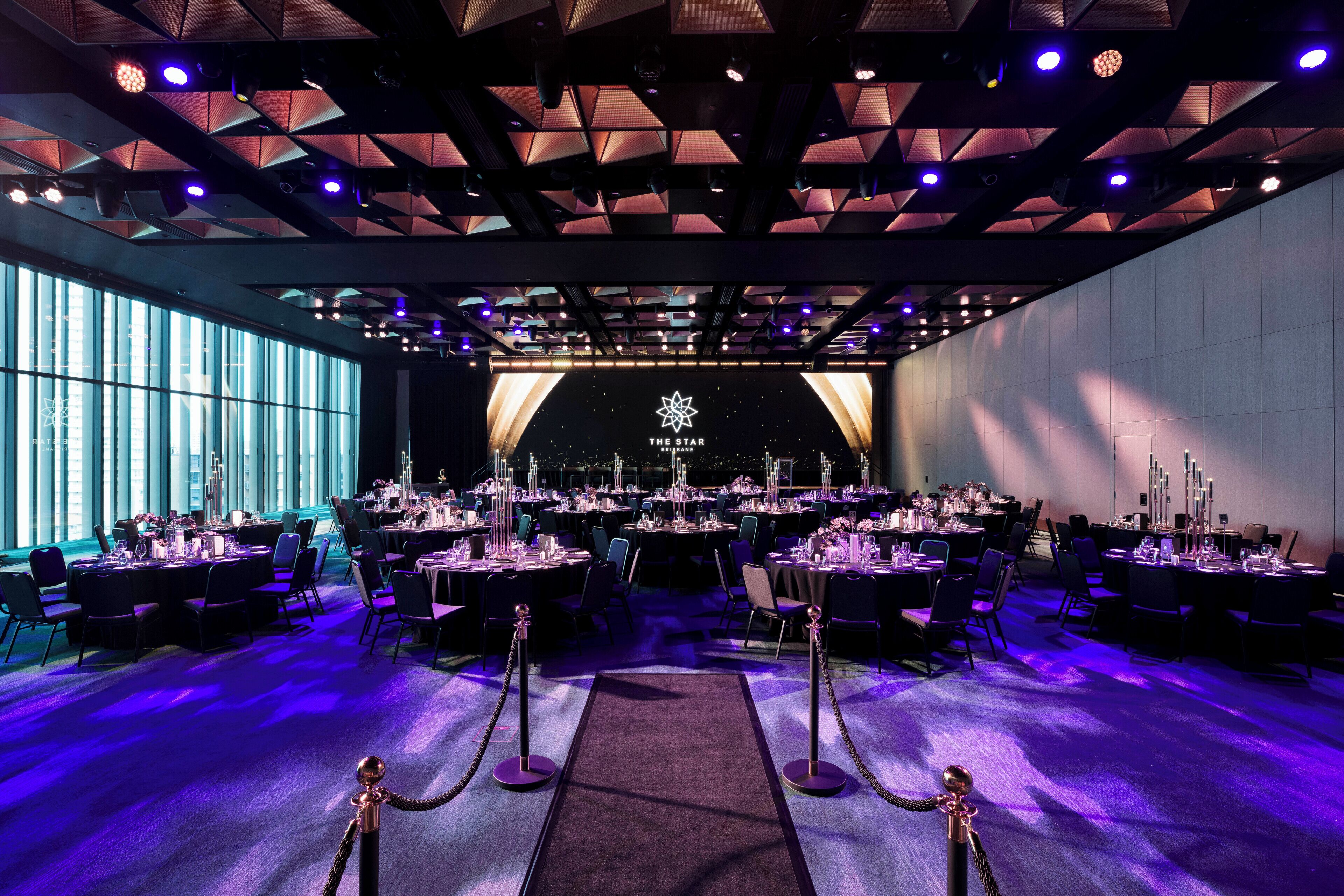 A banquet hall with tables set for a formal event, a stage with a logo, and a view of the city through large windows.