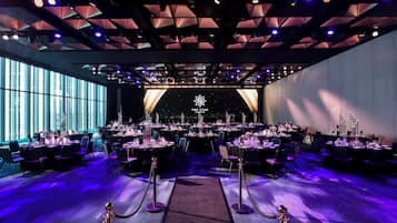 A banquet hall with tables set for a formal event, a stage with a logo, and a view of the city through large windows.